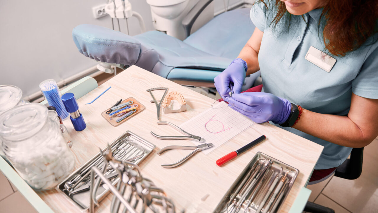 Top view of female dentist in sterile gloves cutting braces wire while sitting at the table with orthodontic instruments and tools. Woman orthodontist preparing wire for braces attachment.